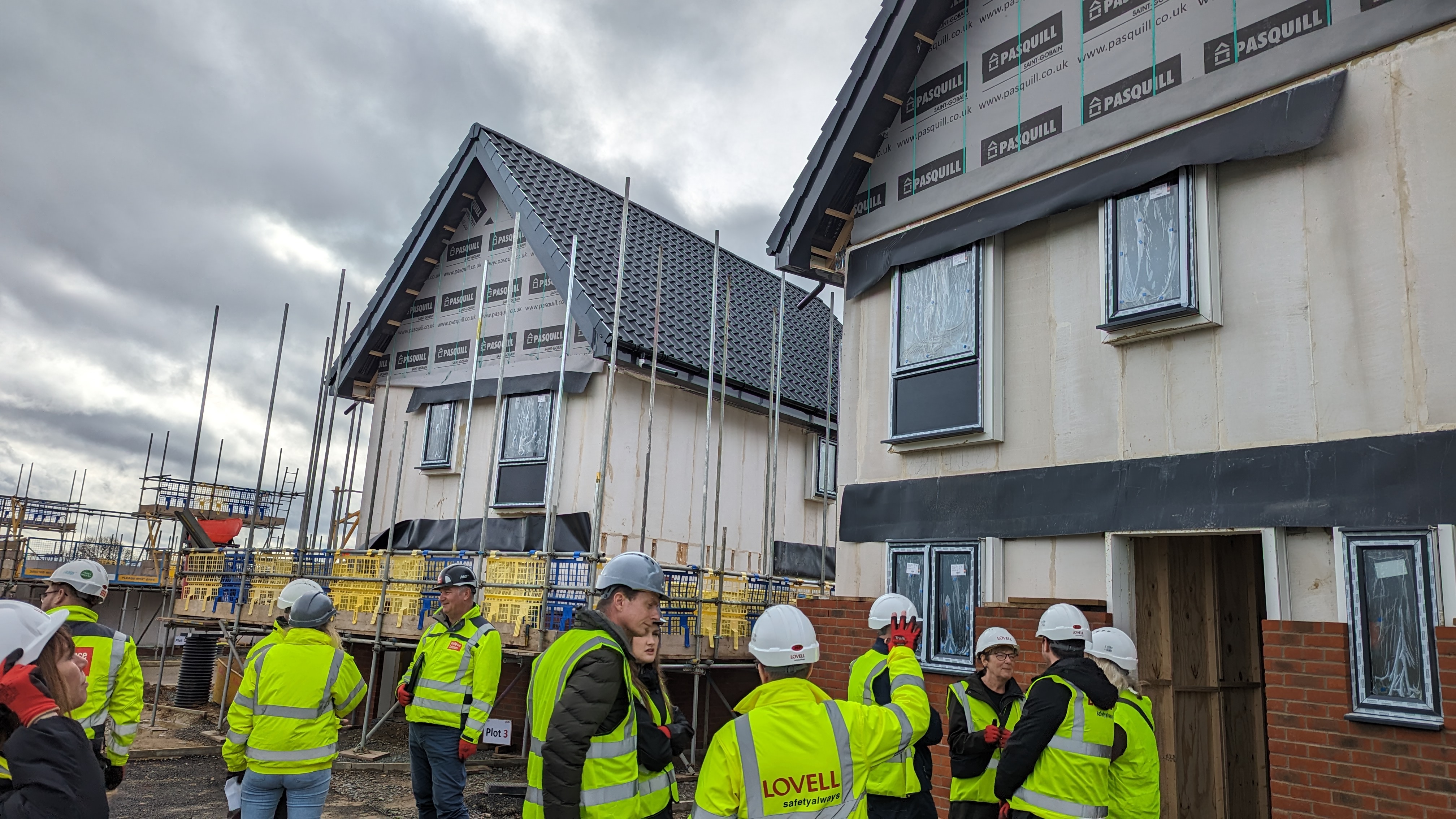 A group of delegates from Flagship Homes, Lovell, and Breckland Council wearing hi-vis vests and hard hats gather outside the in-progress houses on site at Crown View in Great Ellingham.