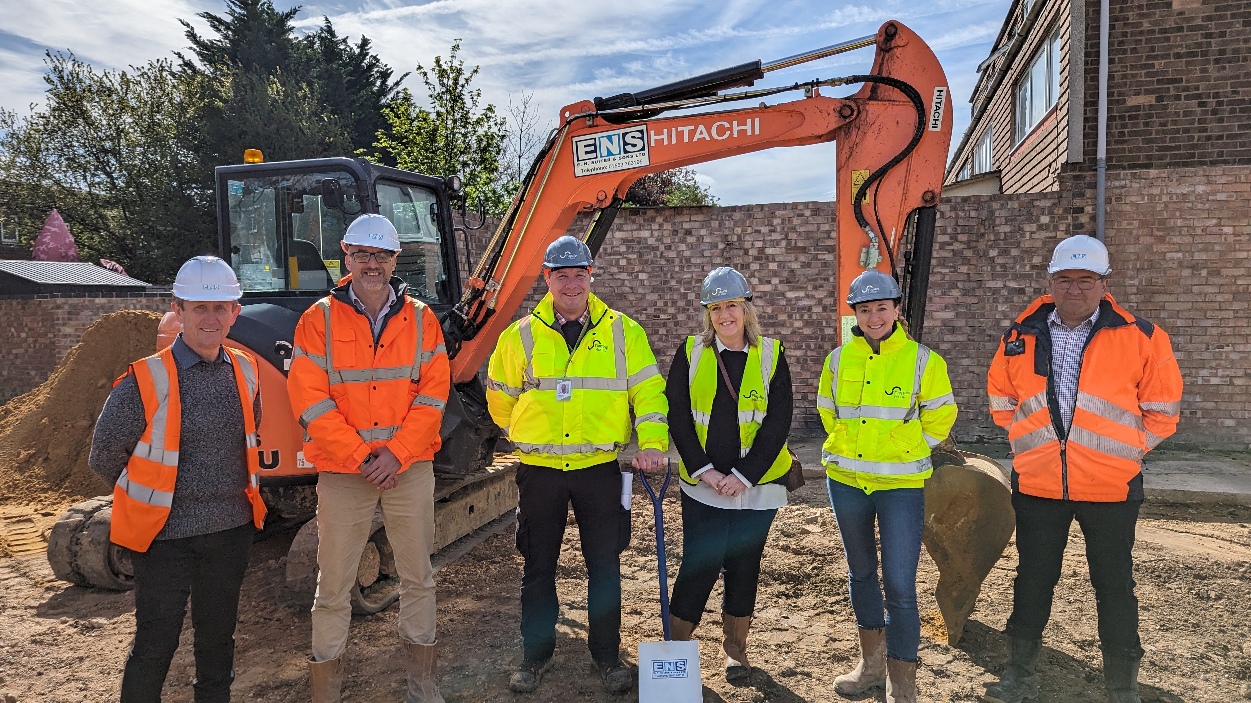 L-R: Martin Kearns, Steven Gould (EN Suiter), Simon Floor, Lisa Davis. Anna Cook-Bacon (Flagship Homes), and Ian Reeve (En Suiter) on one of the garage sites to be turned into new affordable homes in Mildenhall. All 6 are standing in hi-vis jackets in front of a small digger